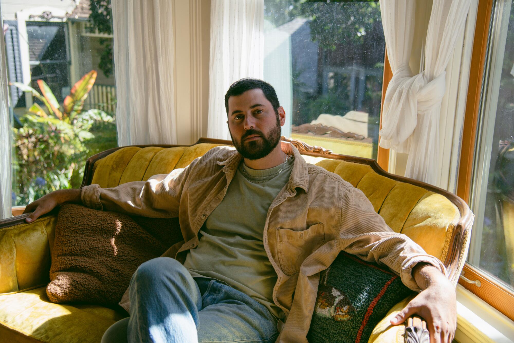 A bearded man in a grey tee shirt, tan jacket and blue jeans sitting on a yellow loveseat with legs crossed.