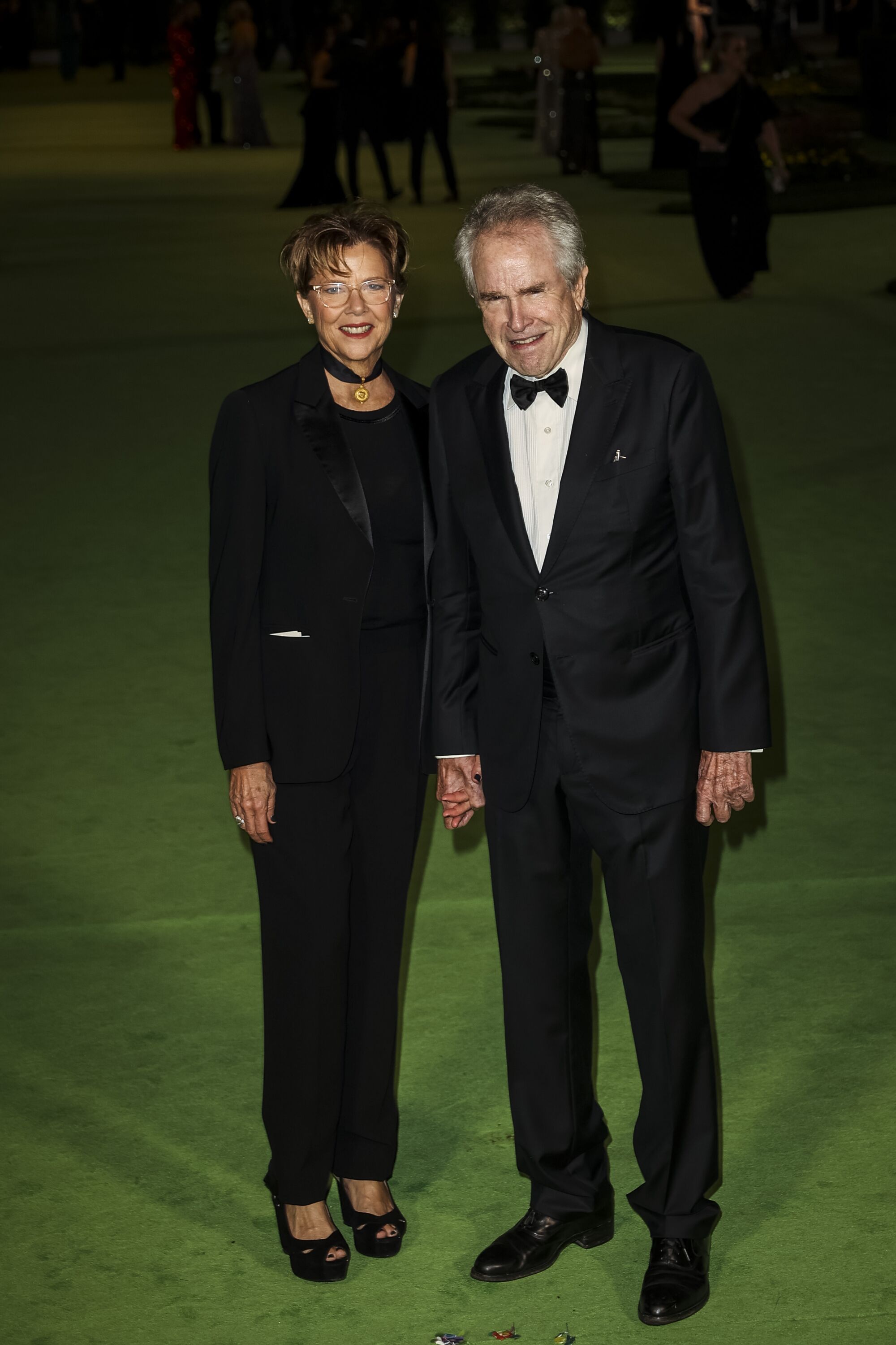 A woman in a black suit and a man in a black tuxedo posing on a green carpet