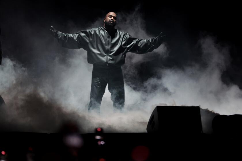 Kanye Wes tin a black jacket, black pants standing on a foggy stage