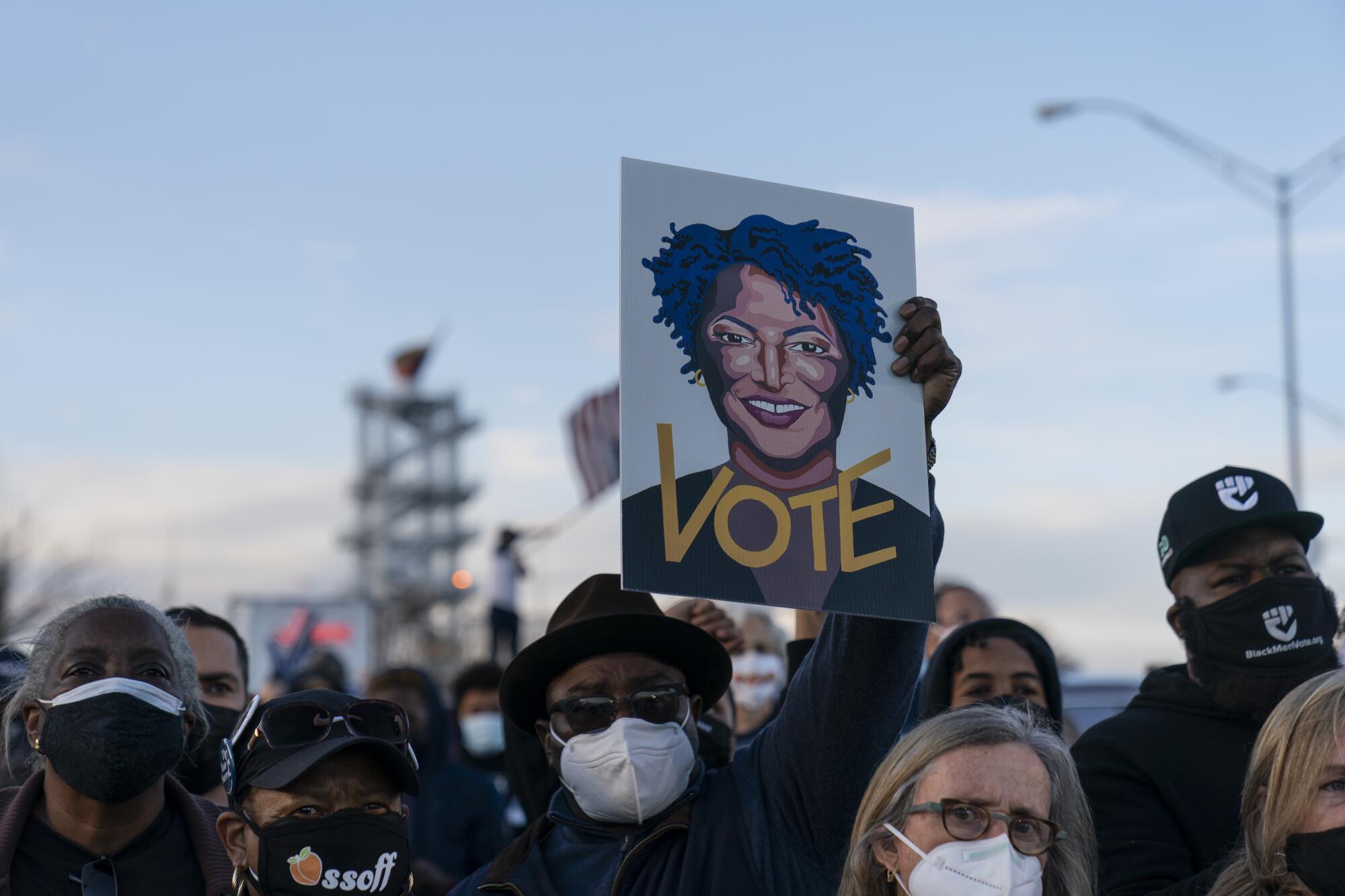 A man in a crowd of people wearing masks holds up an illustration of Stacey Abrams with the word "Vote."