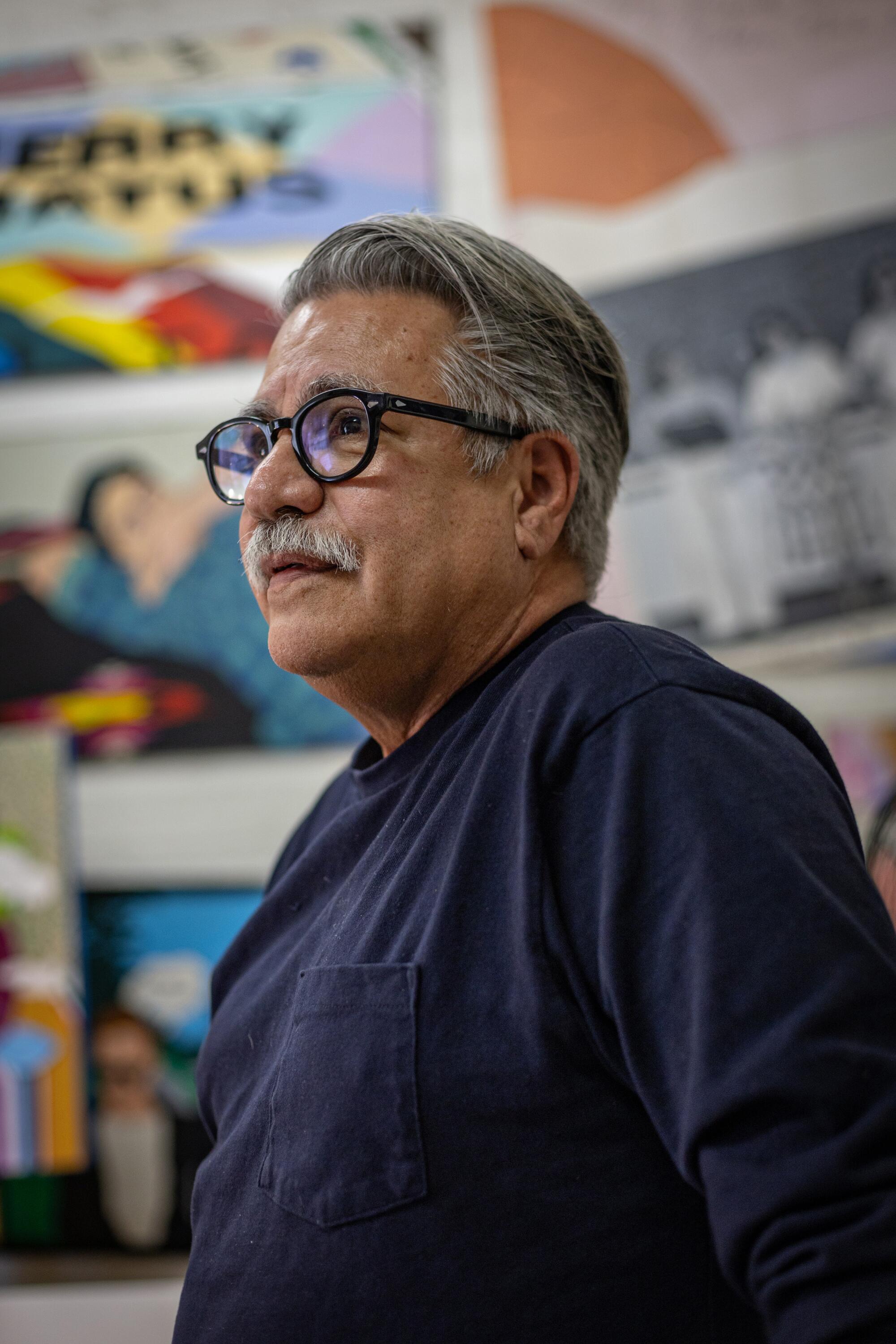 Joey Terrill, a 68-year-old Chicano man wearing a dark shirt, is surrounded by bright paintings in a studio.