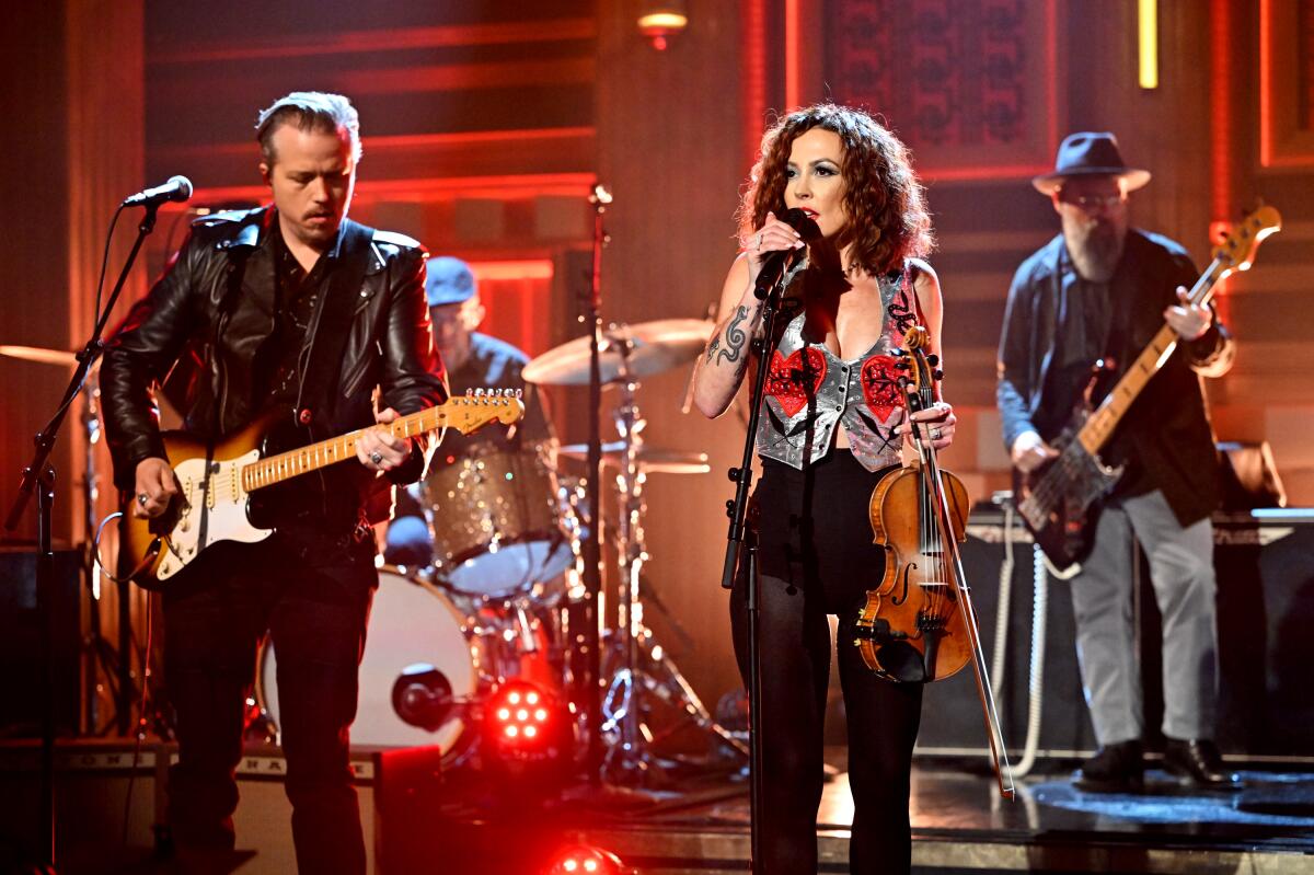 Jason Isbell plays a guitar while Amanda Shires sings into a mic and holds a violin in front of other musicians on stage