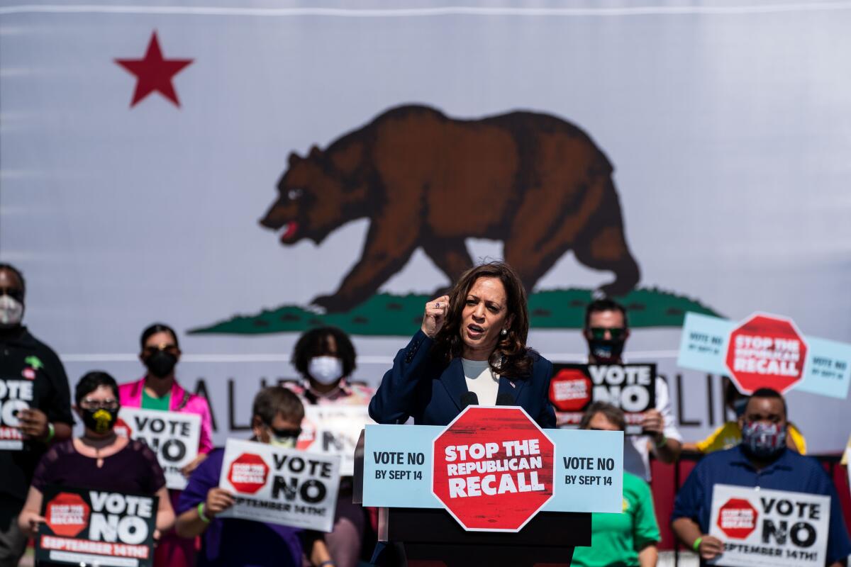 Kamala Harris speaks to the crowd. Behind her is a huge California flag and people holding up anti-recall placards. 