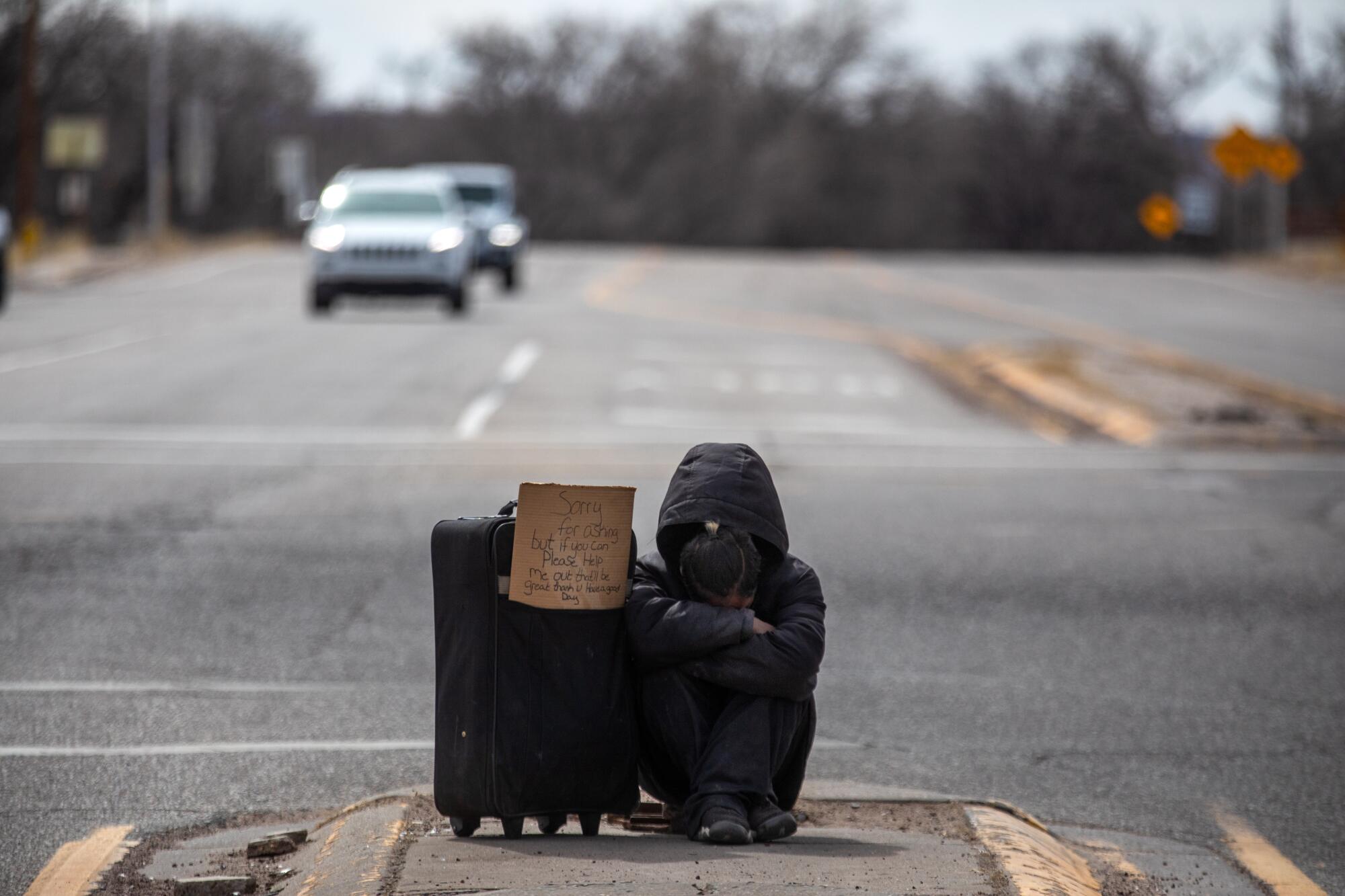 A person sits on the median of the roadway next to a suitcase 