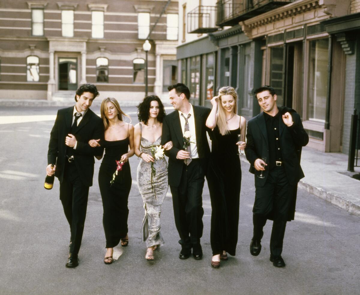 The cast of the sitcom "Friends" walks on a backlot street with gowns on the women and suits on the men