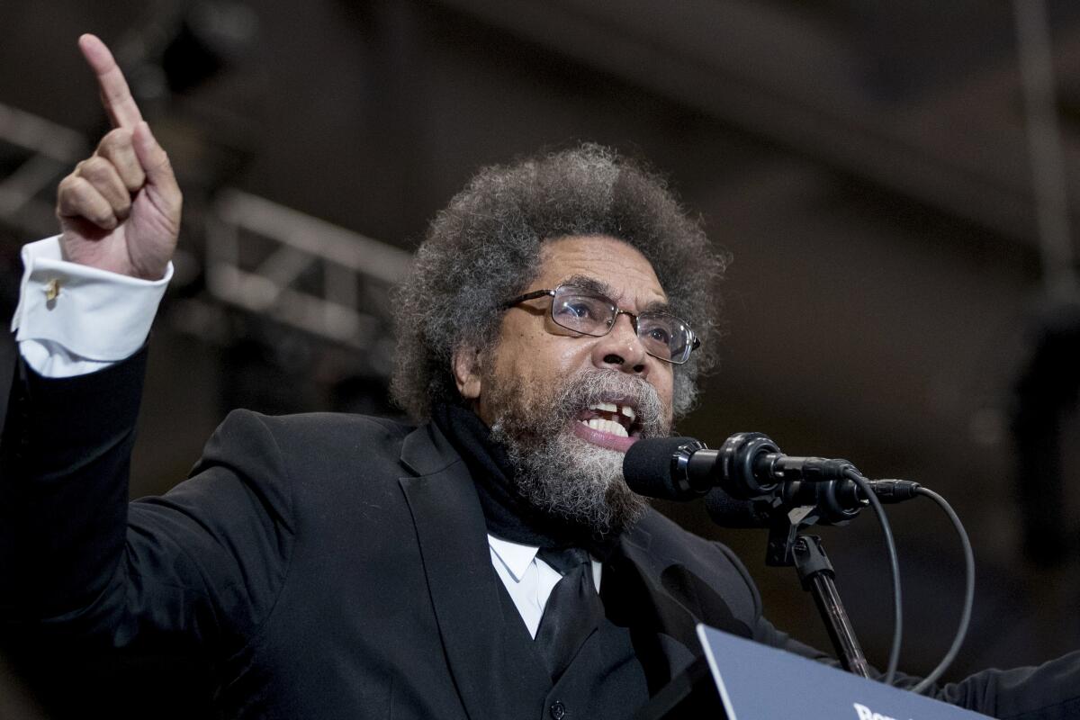 Cornel West pointing as he speaks into microphones on a dark stage