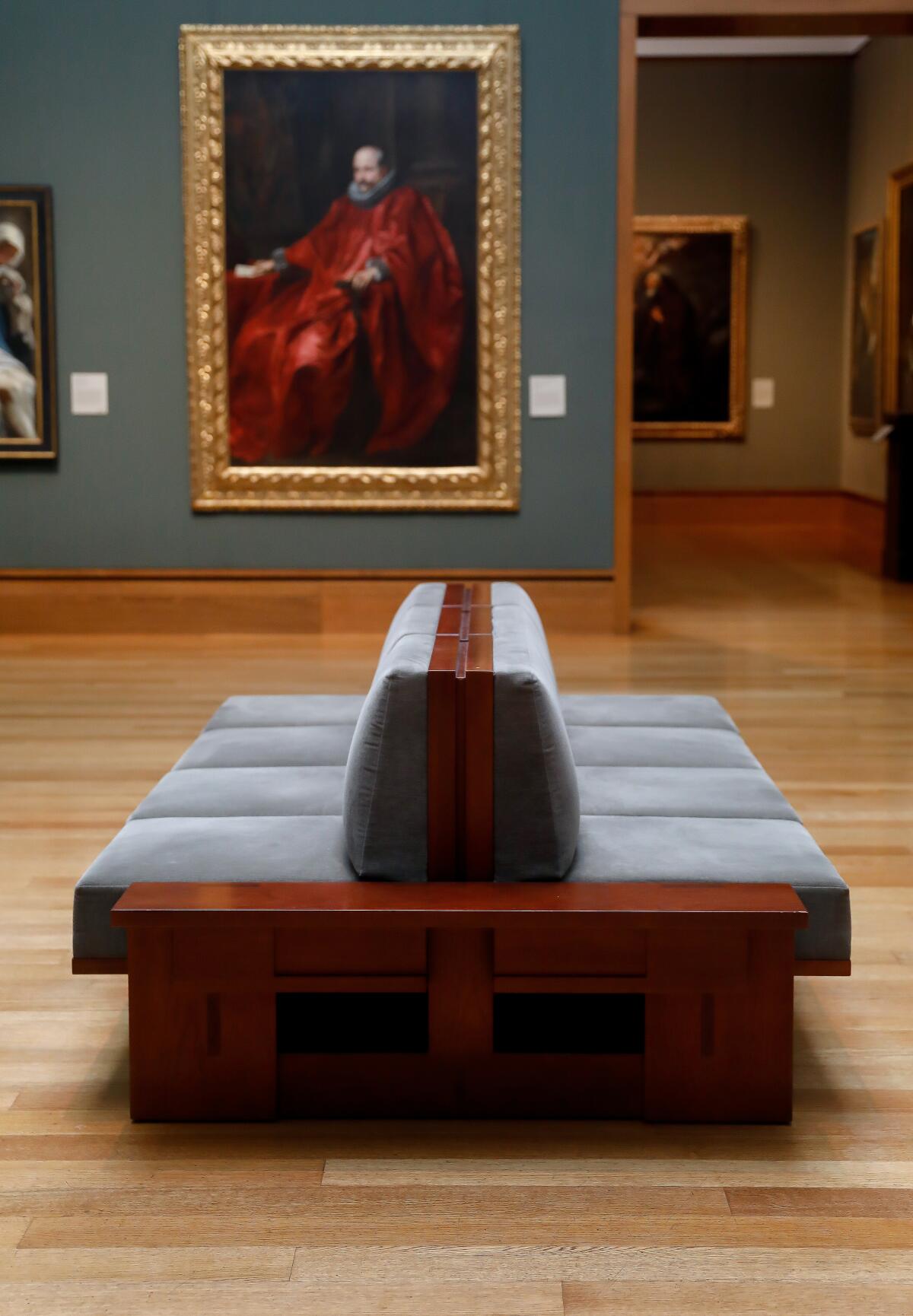 A bench with a hard back and velvety cushions in a gallery with green walls and a portrait of a man in red.