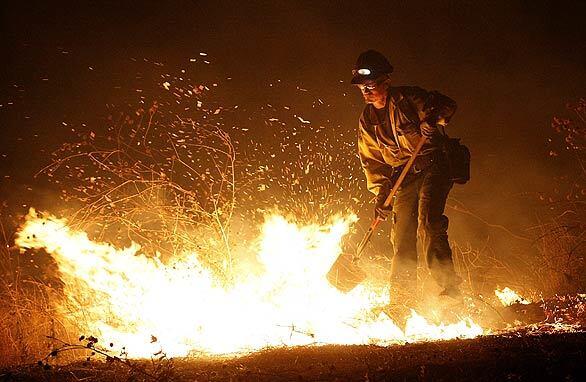 A firefighter beats back flames with a shovel as brush burns around him at a fire fanned by Santa Ana winds in Fontana, San Bernardino County. At least 100 acres have burned. Los Angeles was under a red-flag warning today.