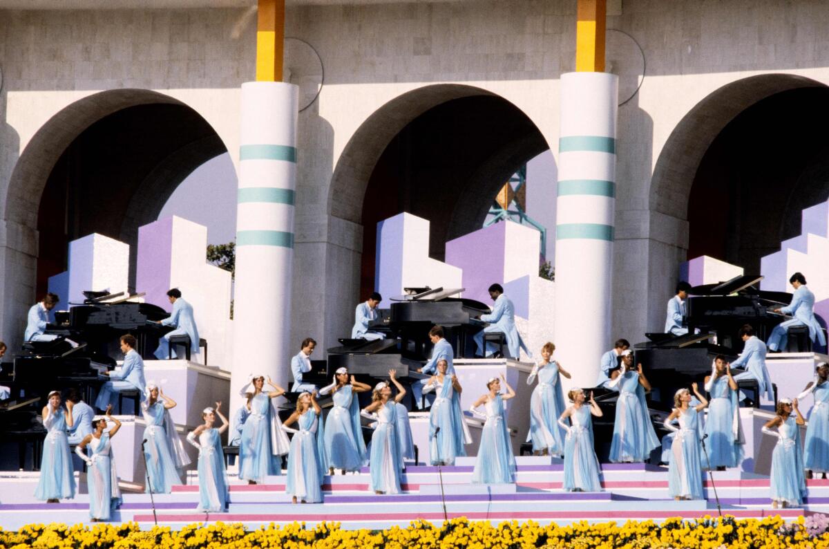Women in light-blue dresses stand and men in light-blue tuxedos play pianos on a large outdoors stage.