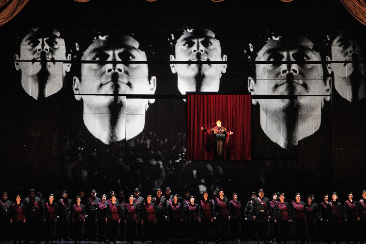 Four faces projected big on a stage with many opera singers below 