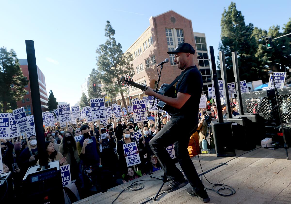 A singer-guitarist performs onstage in front of a crowd holding signs reading: "UAW on strike unfair labor practice."