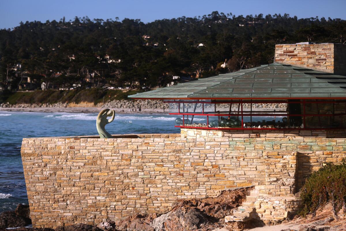 An oceanfront house designed by Frank Lloyd Wright has a green shingled roof, glass windows and a rock wall.