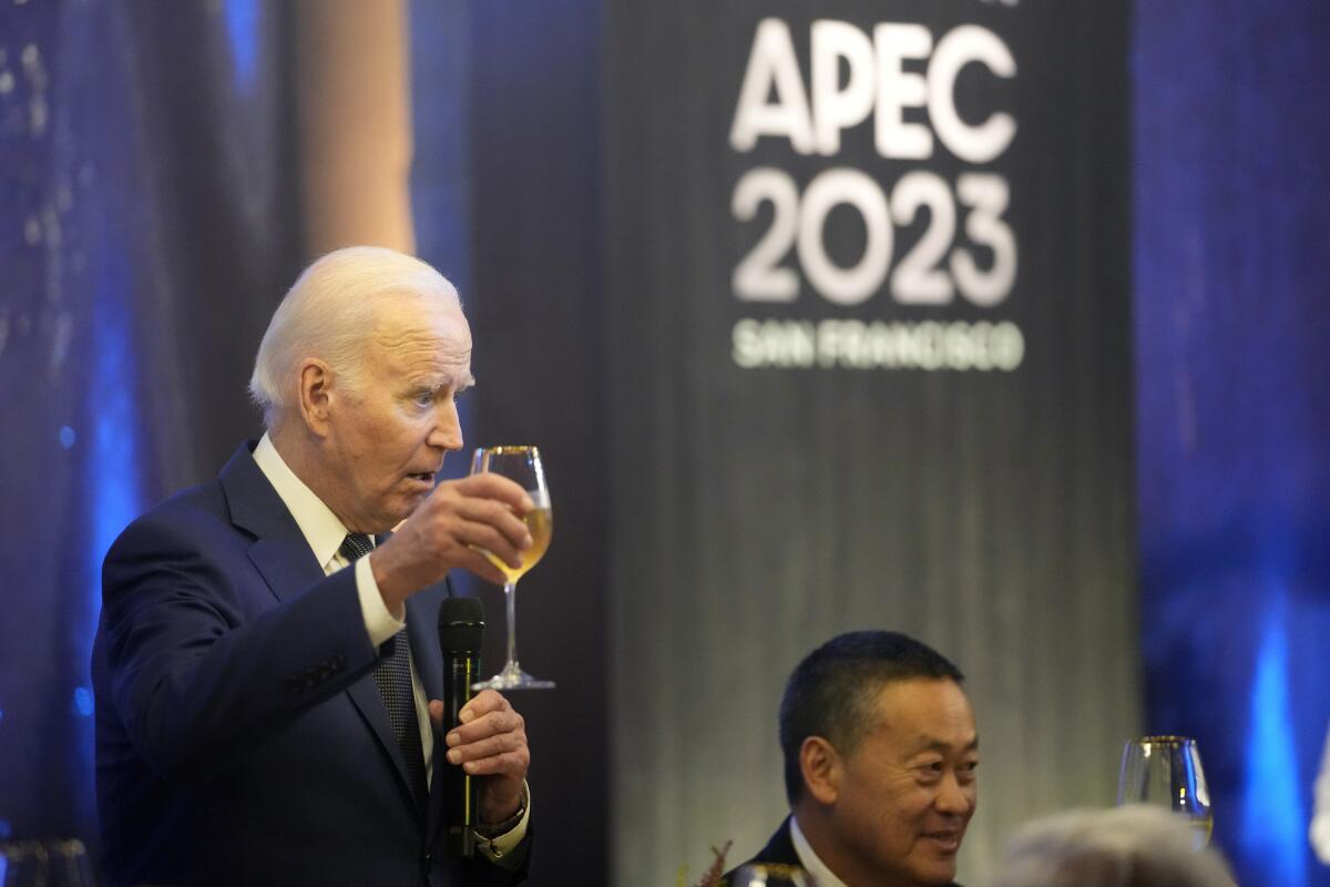 President Biden stands and raises a wine glass while speaking into a microphone near a sign saying APEC 2023 San Francisco.