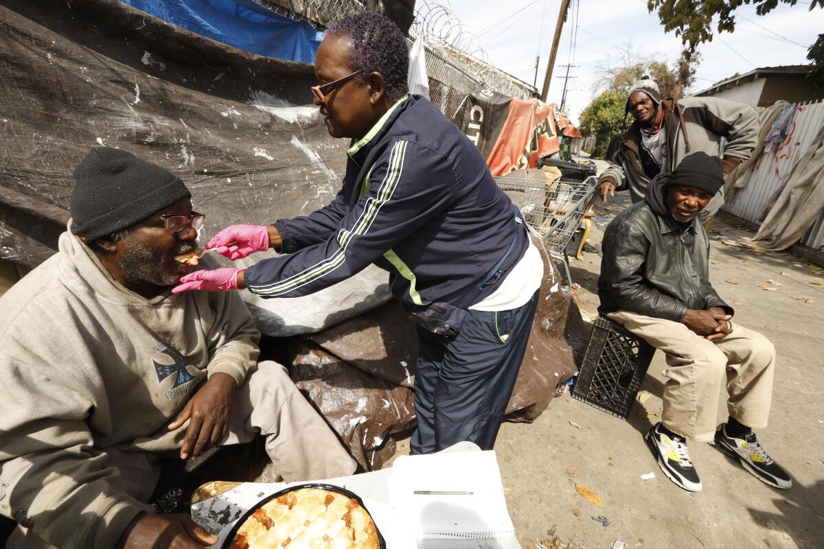A woman feeds pie to a man sitting outside a homeless encampment