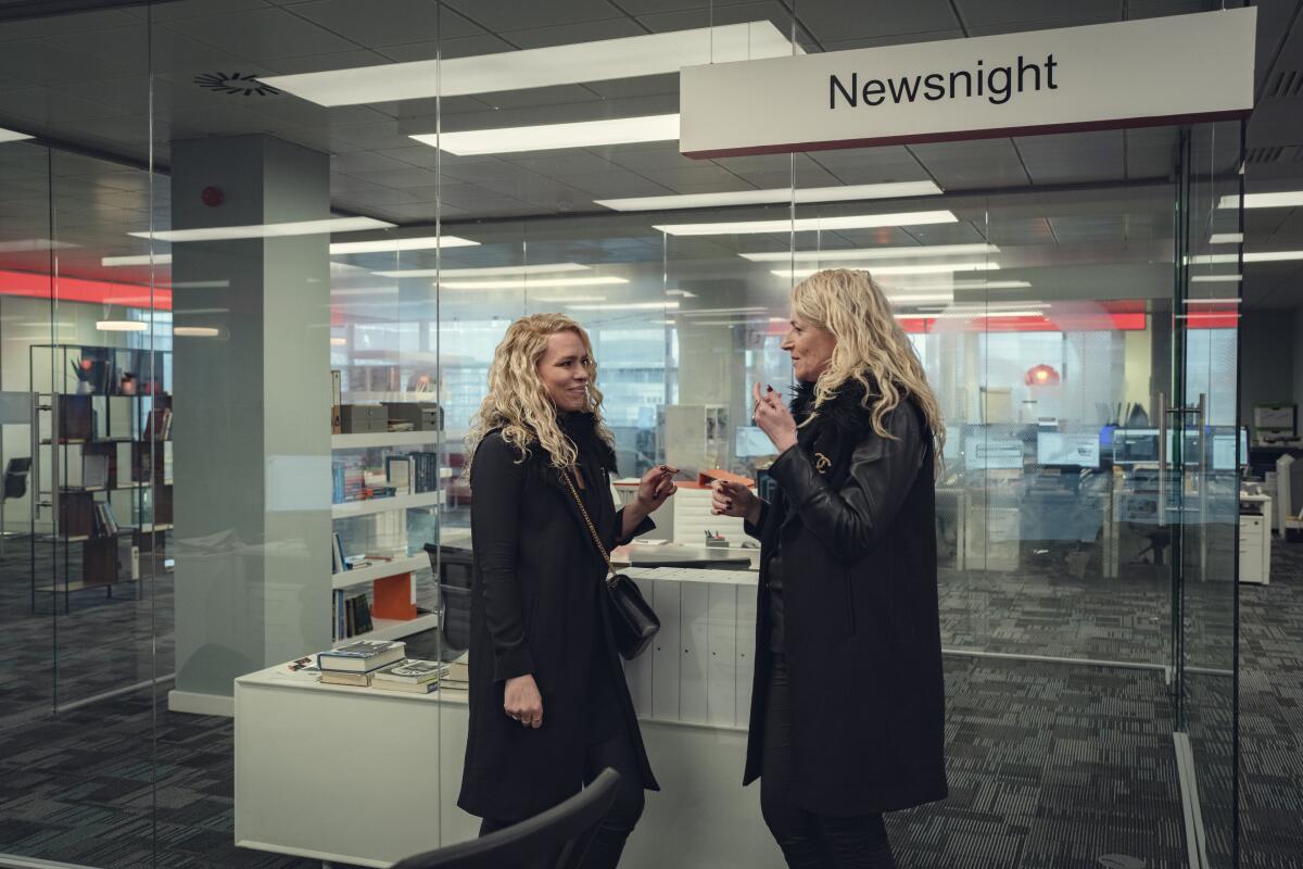 Two blond women in black coats standing in an office chatting.