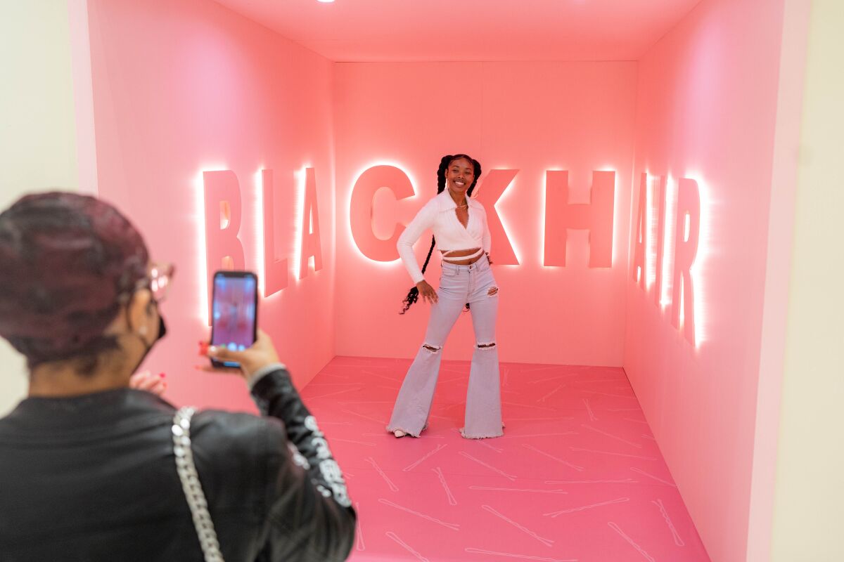 A woman poses for a photo inside a pink room with the words 'Black Hair' on the walls.