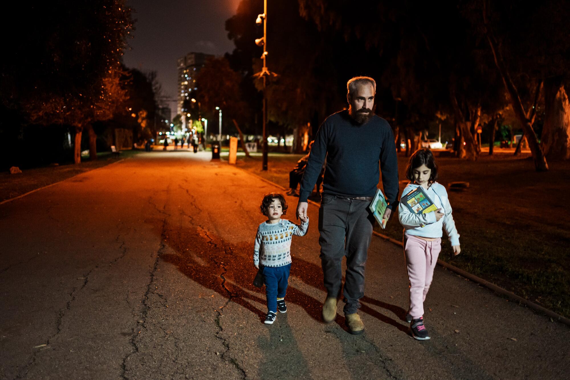 A bearded man holds a young boy's hand while walking next to a young girl, holding a book, on a lit path at night