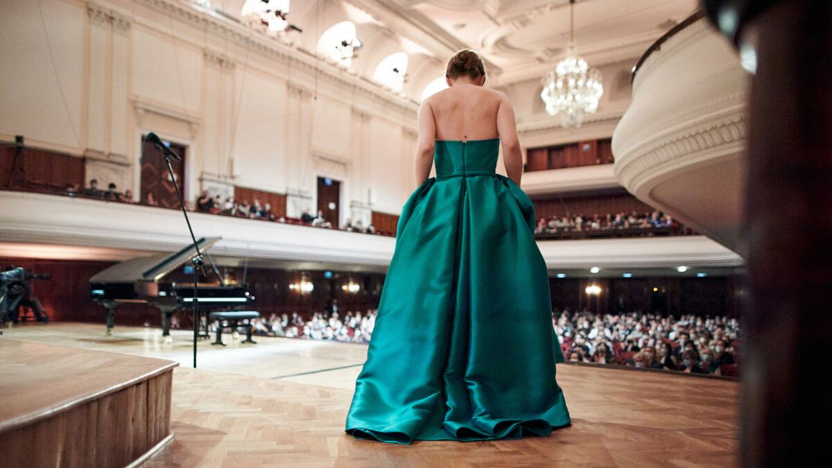 A woman in a gown, standing near a grand piano, faces an audience.