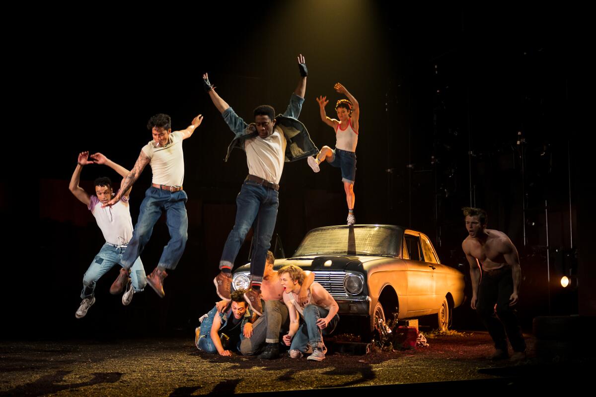 four men jump off the hood of a car while two kneel in front of the car, all in jeans and a white t-shirt 