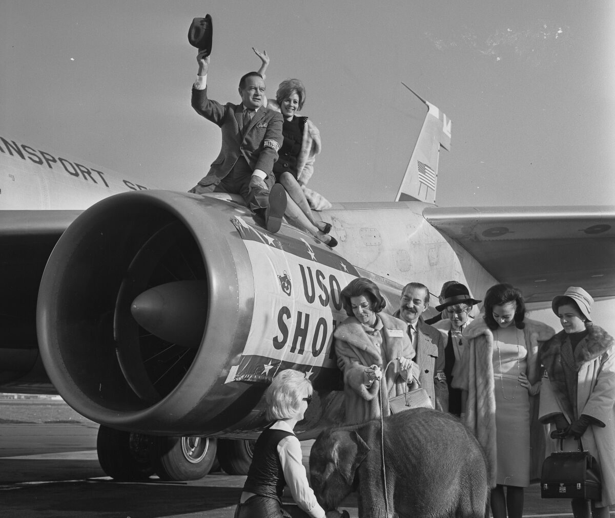 A man waves a fedora while atop a jet engine with a waving woman. Several people stand below with a baby elephant.