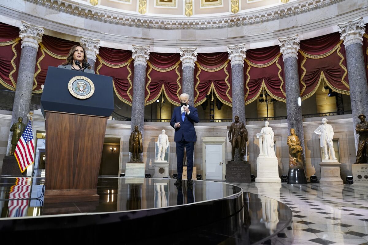Kamala Harris speaks at a podium in a rotunda filled with statues; President Biden stands nearby.