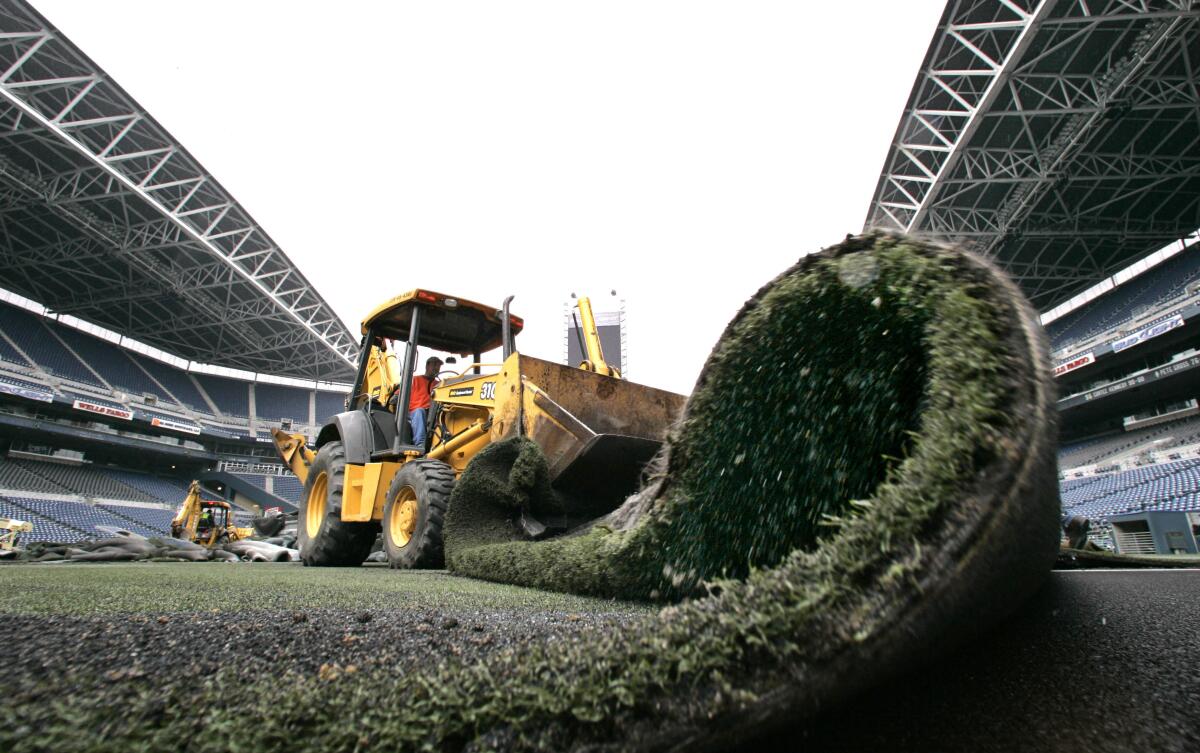 A worker using a construction vehicle to remove artificial turf from a stadium field.