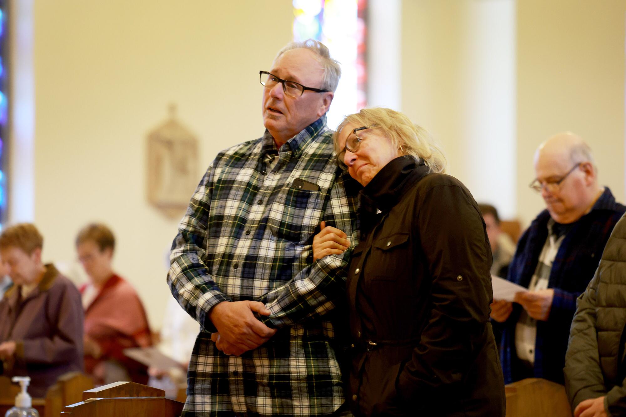 A woman rests her head on the shoulder of a man and holds his arm as they stand in the pews of a church