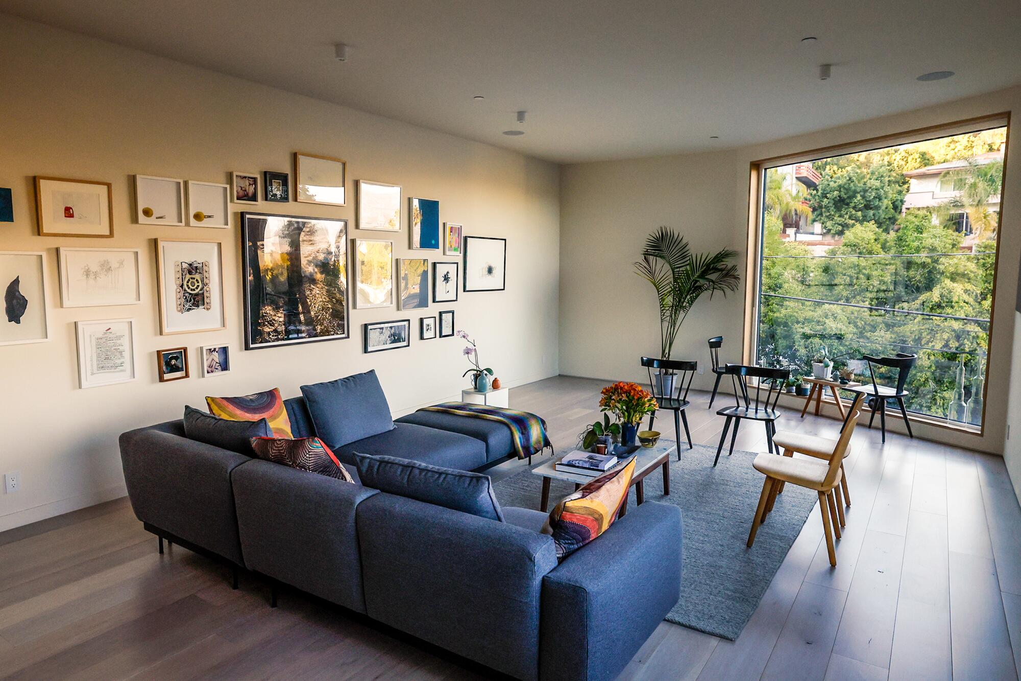 A blue couch sits in the living room near a wall covered in frames and a large window with a view of a green hillside. 