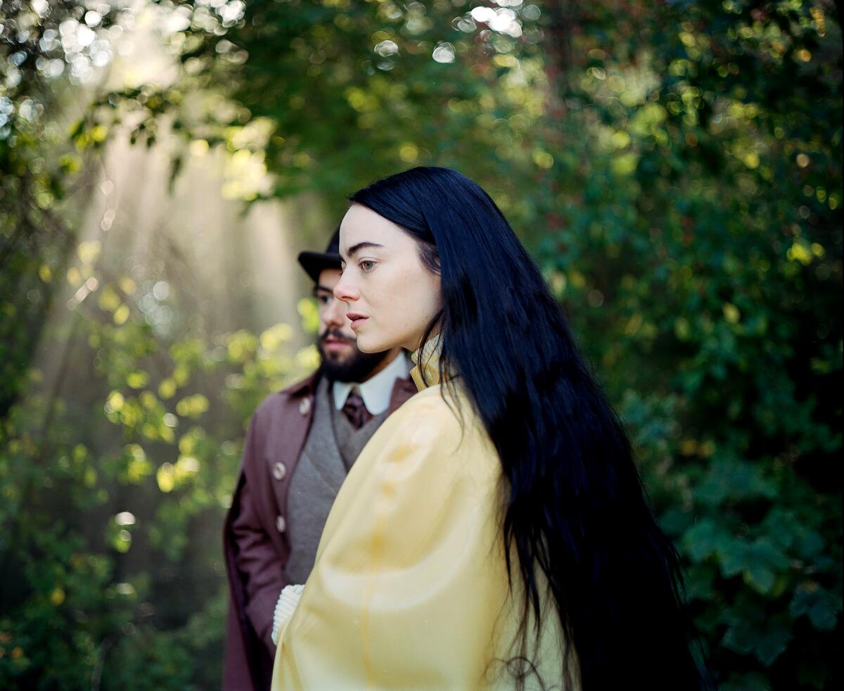 A suited man in a hat and a woman with long dark hair stroll outdoors.