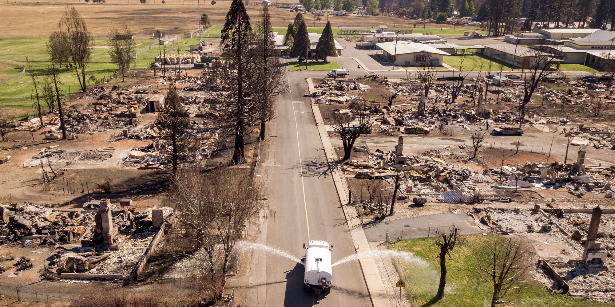 A water truck sprays both sides of a fire-ravaged street