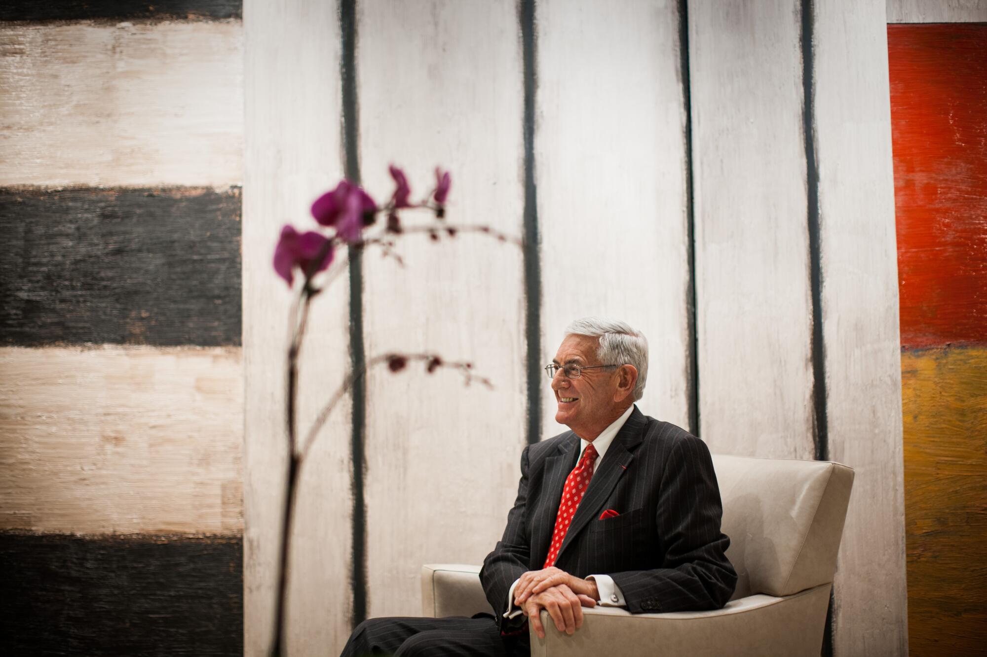 Eli Broad sits in an armchair in front of an oversized painting with black and white stripes.