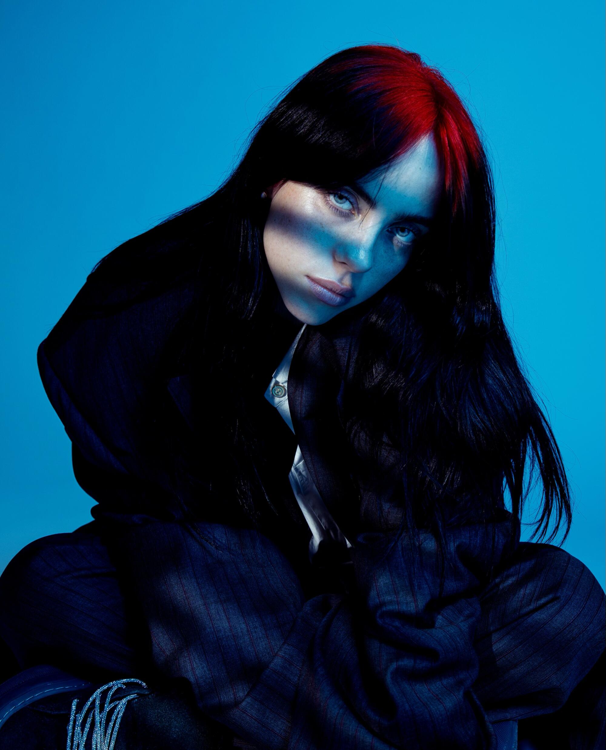 Billie Eilish, with red streaks in her hair, poses in front of a blue backdrop. 