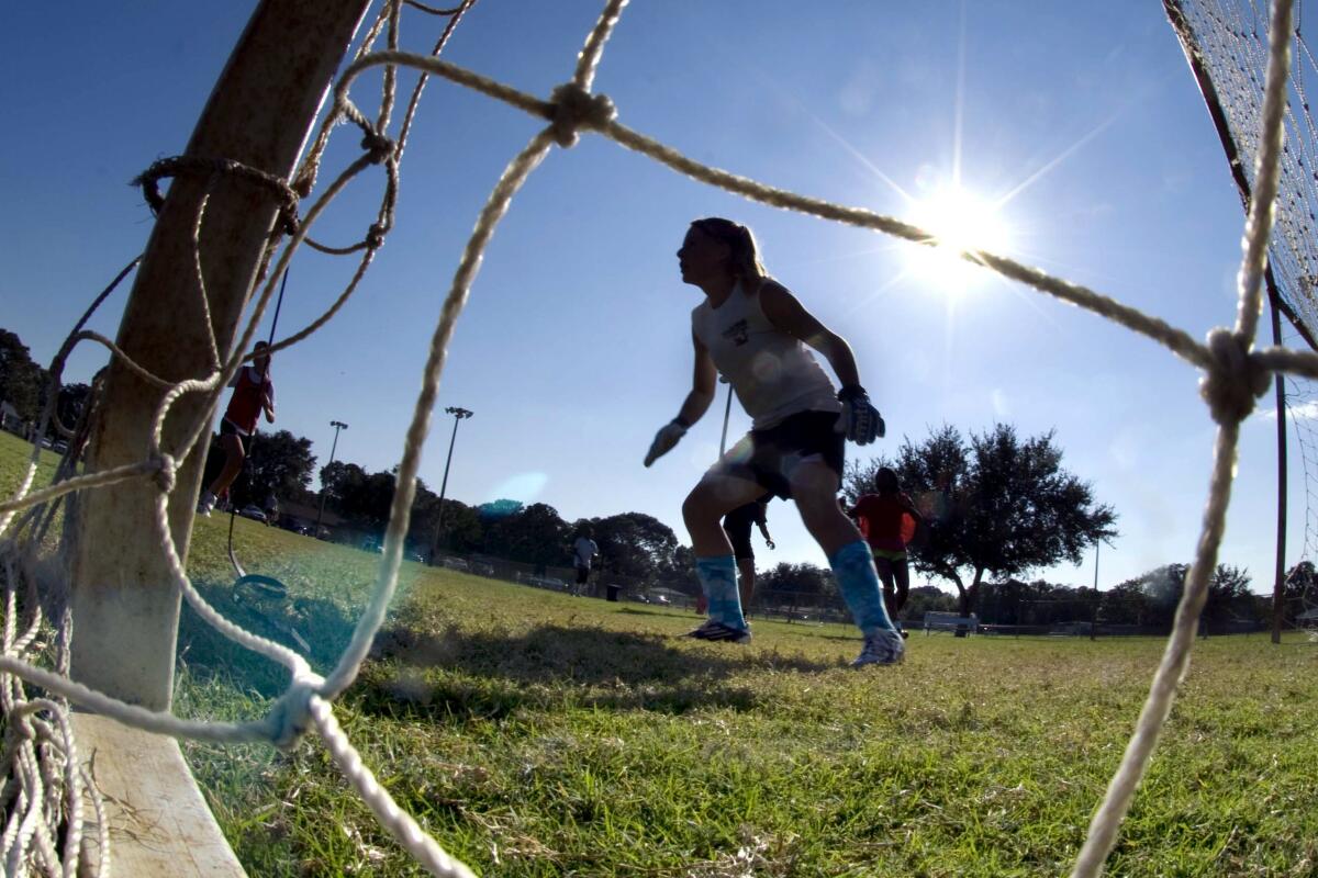 A woman stands on a soccer field, as seen through goal netting. 