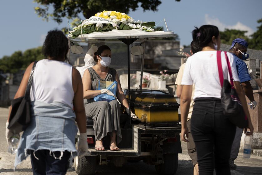 A woman wearing a face mask sits next the coffin of a relative as she's transported by a cemetery worker to the burial site at the Nossa Senhora das Gracas cemetery in Duque de Caxias, Rio de Janeiro, Brazil, Monday, April 27, 2020. The deceased person was previously held in a refrigerator for confirmed and suspected victims of of COVID-19, according to the administration of the cemetery. (AP Photo/Silvia Izquierdo)