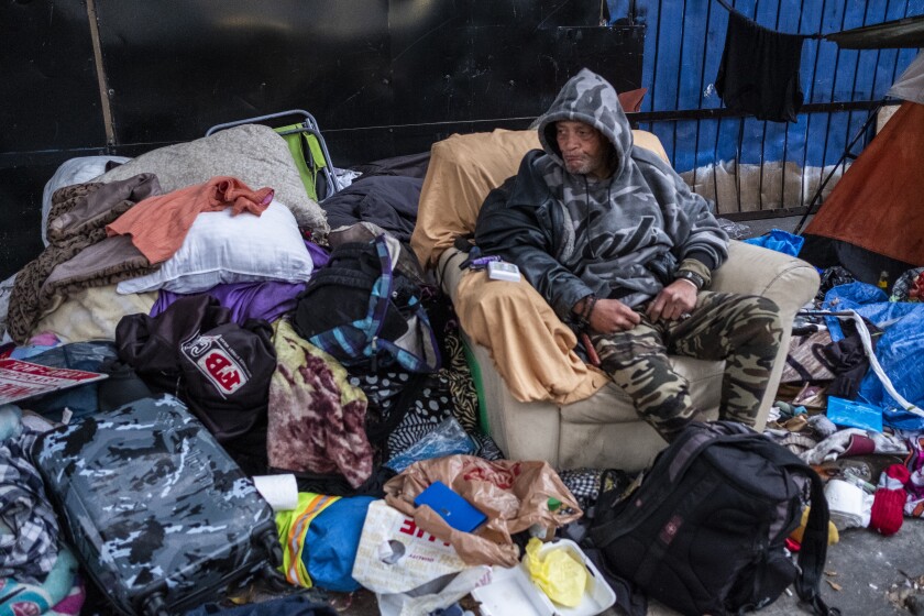 A man sits in a chair next to mounds of clothes on the sidewalk.