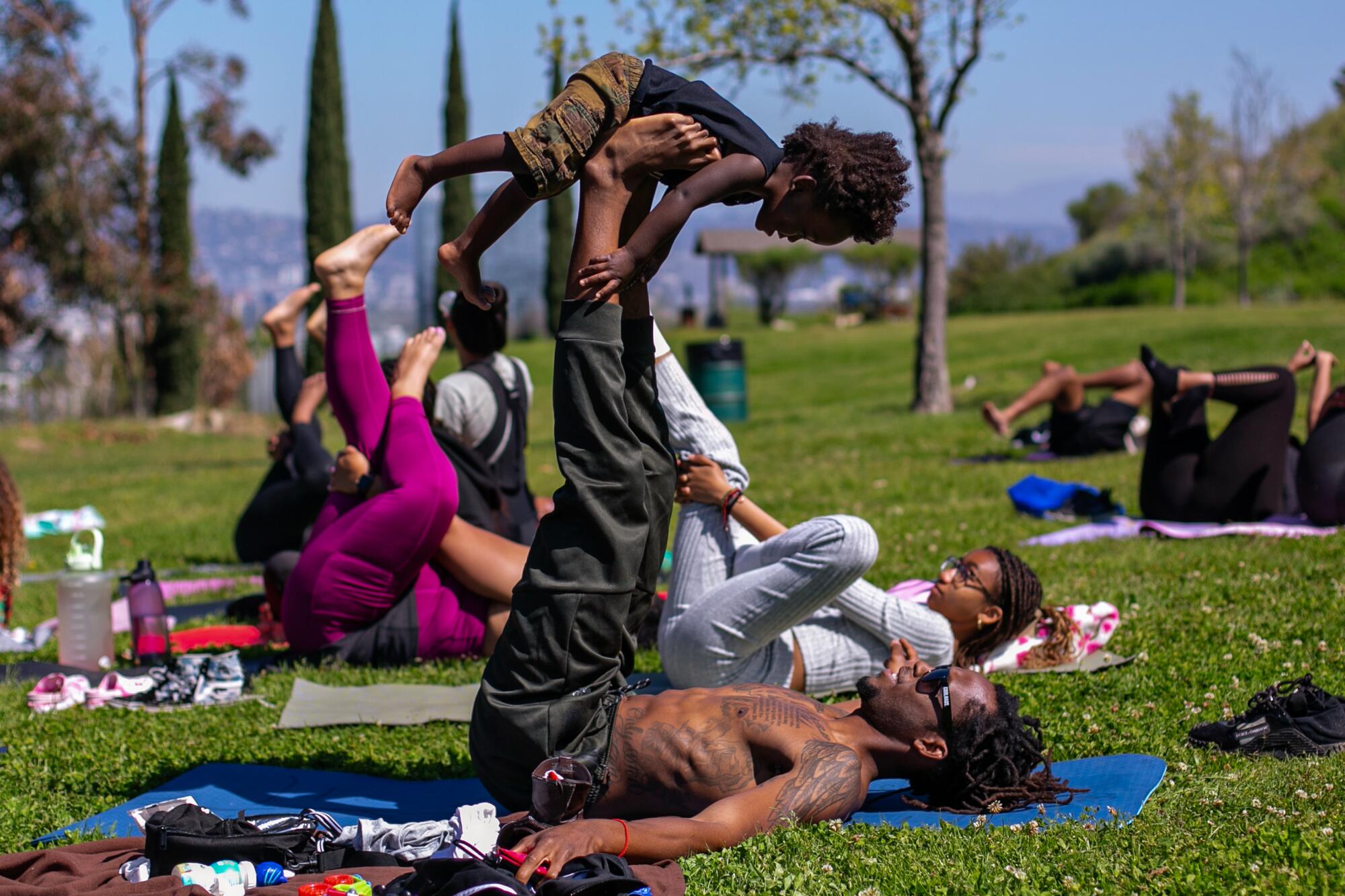 Several people lie on mats and raise their legs up. One man lifts a child on his raised feet.