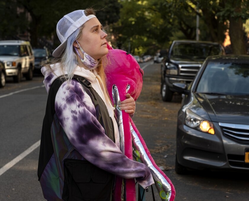A woman in a tie-dyed hoodie and backward cap stands on a street.