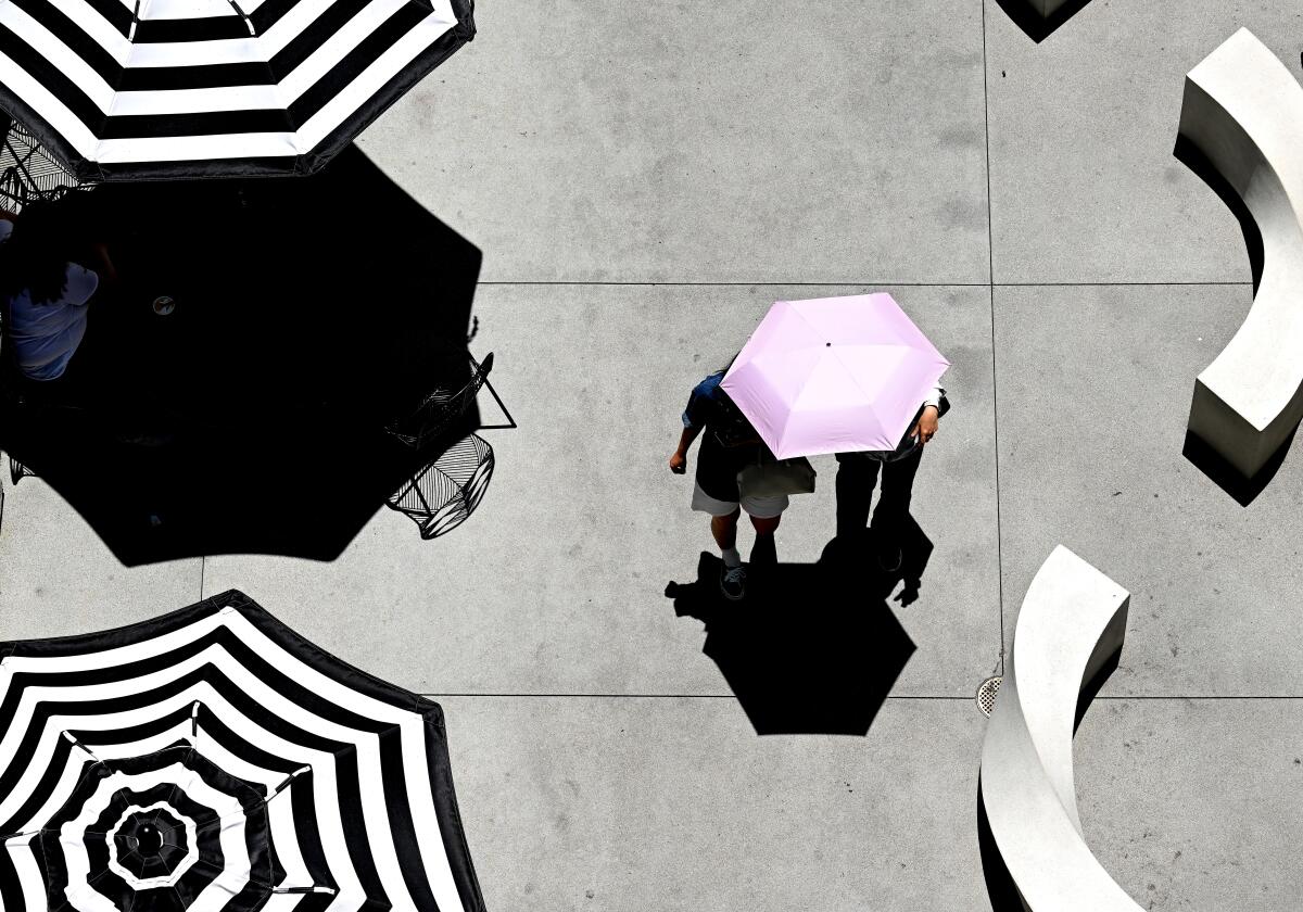 Two people, seen from above, walk with a pink umbrella past black and white striped patio umbrellas 
