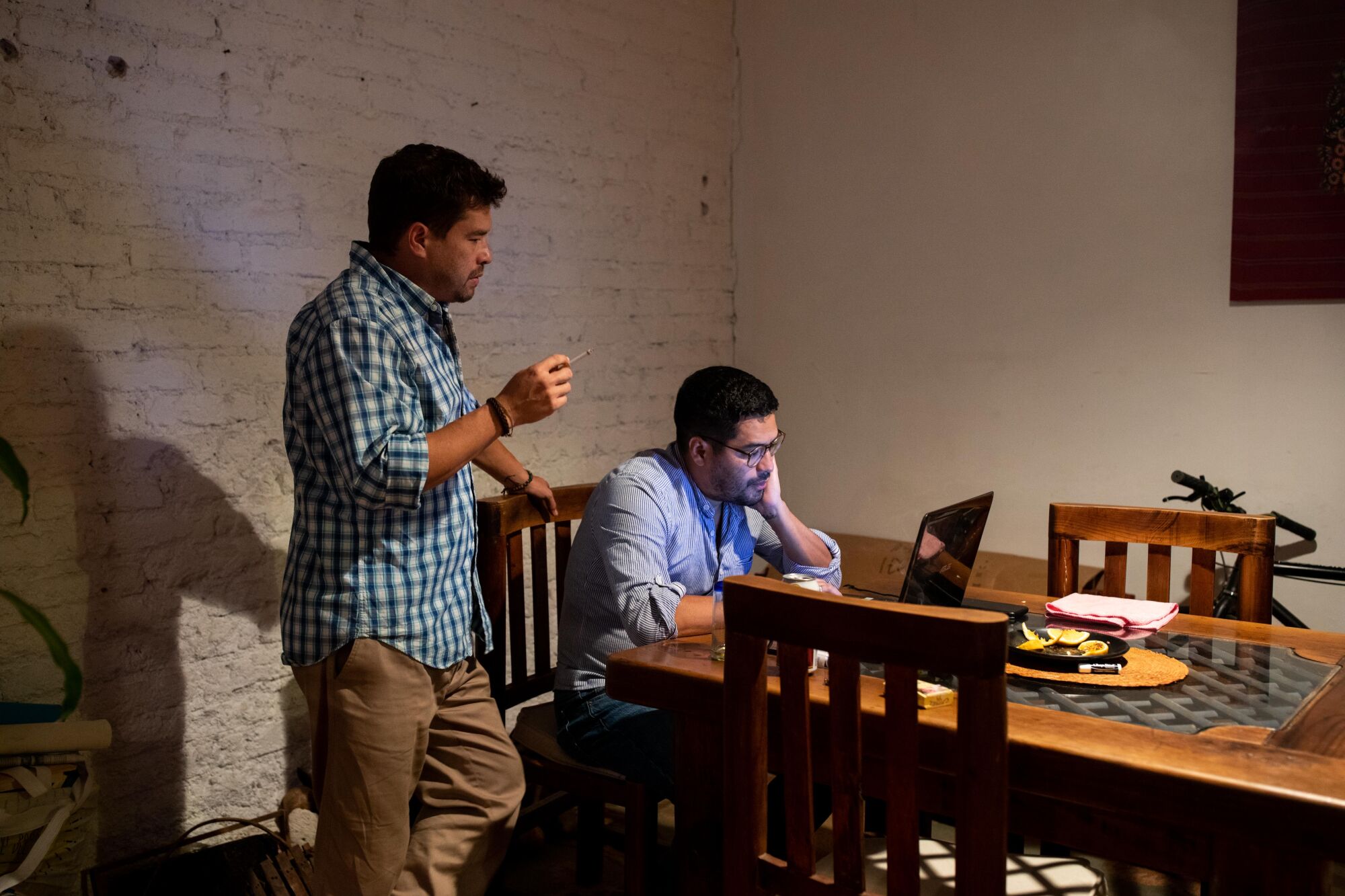 A man sits looking at a laptop on a large table as another man stands next to him. 