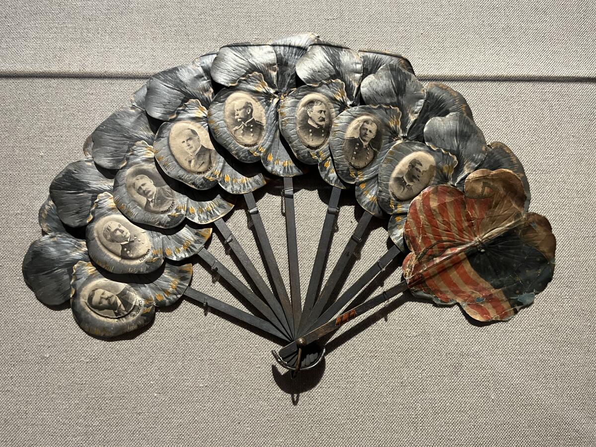 A vintage metal hand fan in a museum vitrine features a U.S. flag and black-and-white images of military leaders.