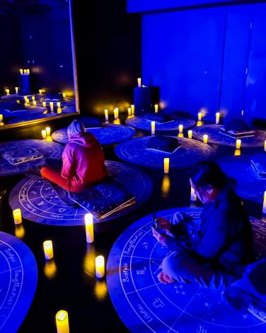 People sit cross-legged on round mats in a dark room illuminated with electric candles.