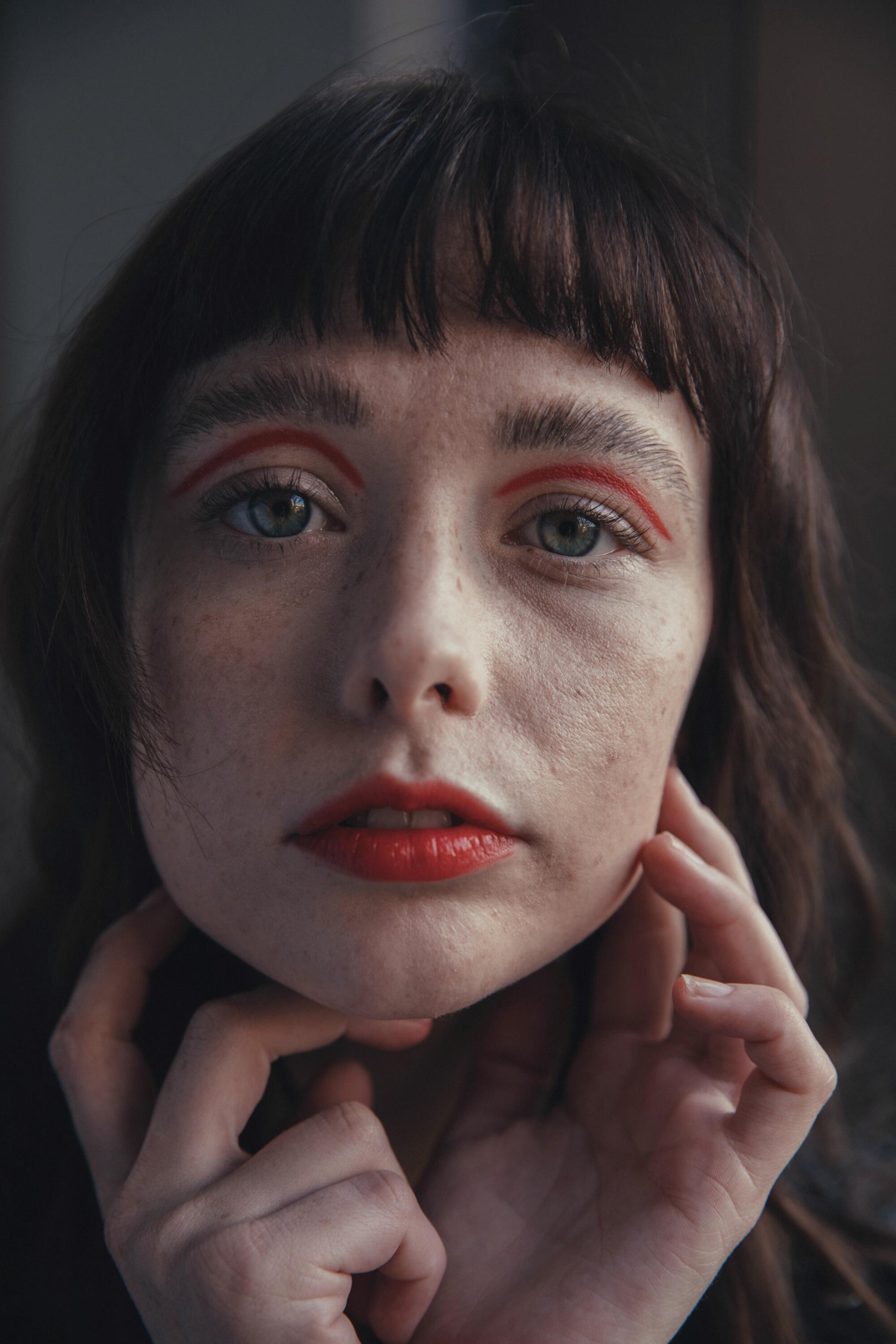 A woman with red lipstick and red eye makeup holds her hands to her face.