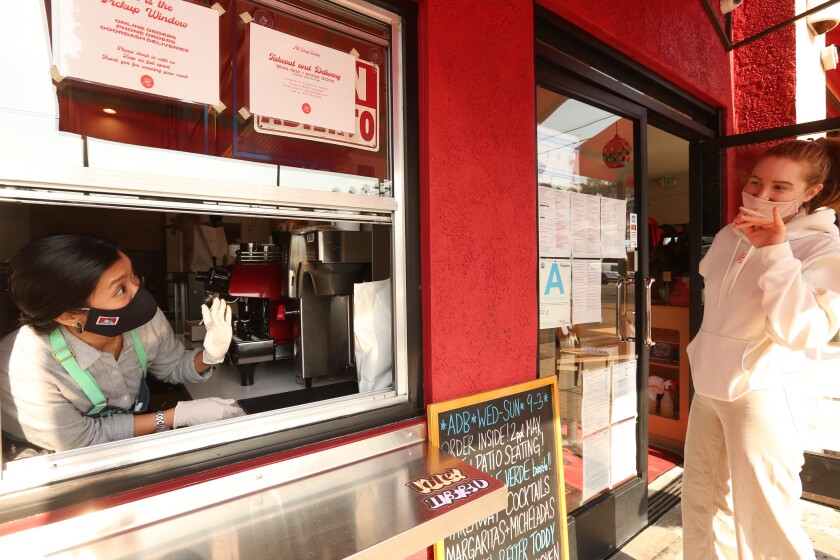 A restaurant owner in a mask and gloves leans out of the takeout window of her restaurant