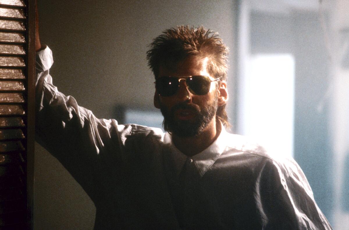 A man in the 1980s with sunglasses and a mullet and beard leaning against a wall
