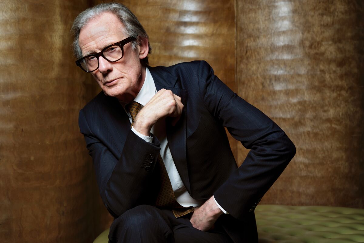 Bill Nighy sits on a bench and leans against a wall for a portrait.