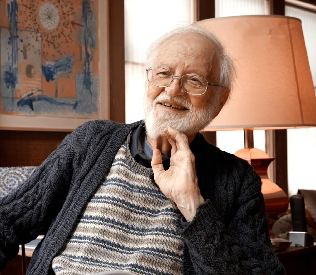 Jim Melchert, an older man with a beard, sits in a chair before a lamp wearing a striped sweater. 