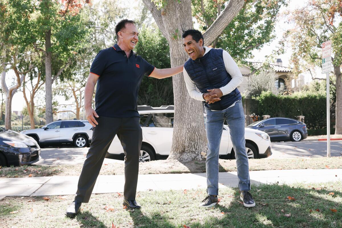 Two men laugh while standing under a tree near a sidewalk
