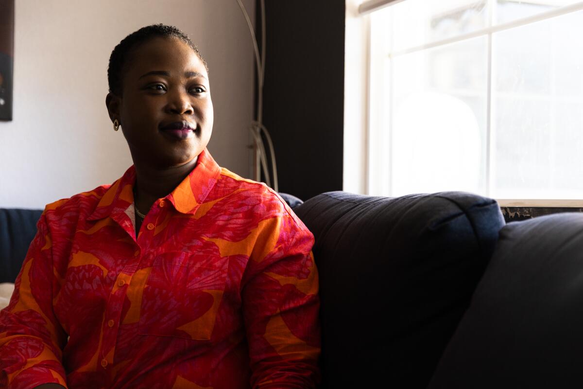 A woman in a bright orange and red shirt sits on a couch in from of a window