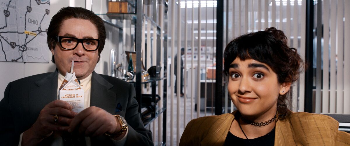 A woman sits in an office with a man drinking milk from a carton.