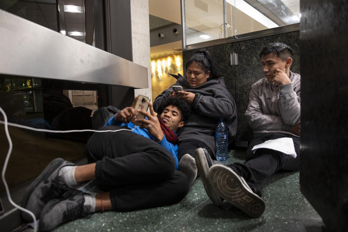 Three people relaxing on a floor, one man resting his head on a seated woman's lap as both look at their phones.